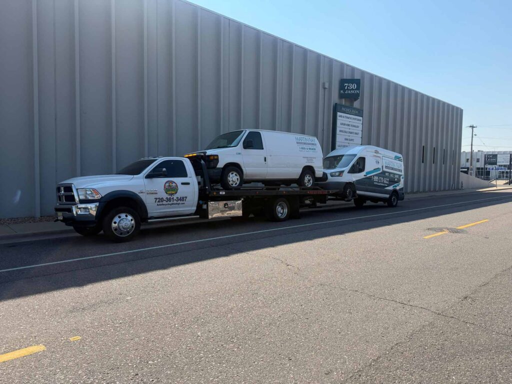 White Auto Recycling Mile High Ram 5500 flatbed tow truck carrying a white Ford cargo van and towing a second Ford Transit van outside 730 South Jason Street in Denver