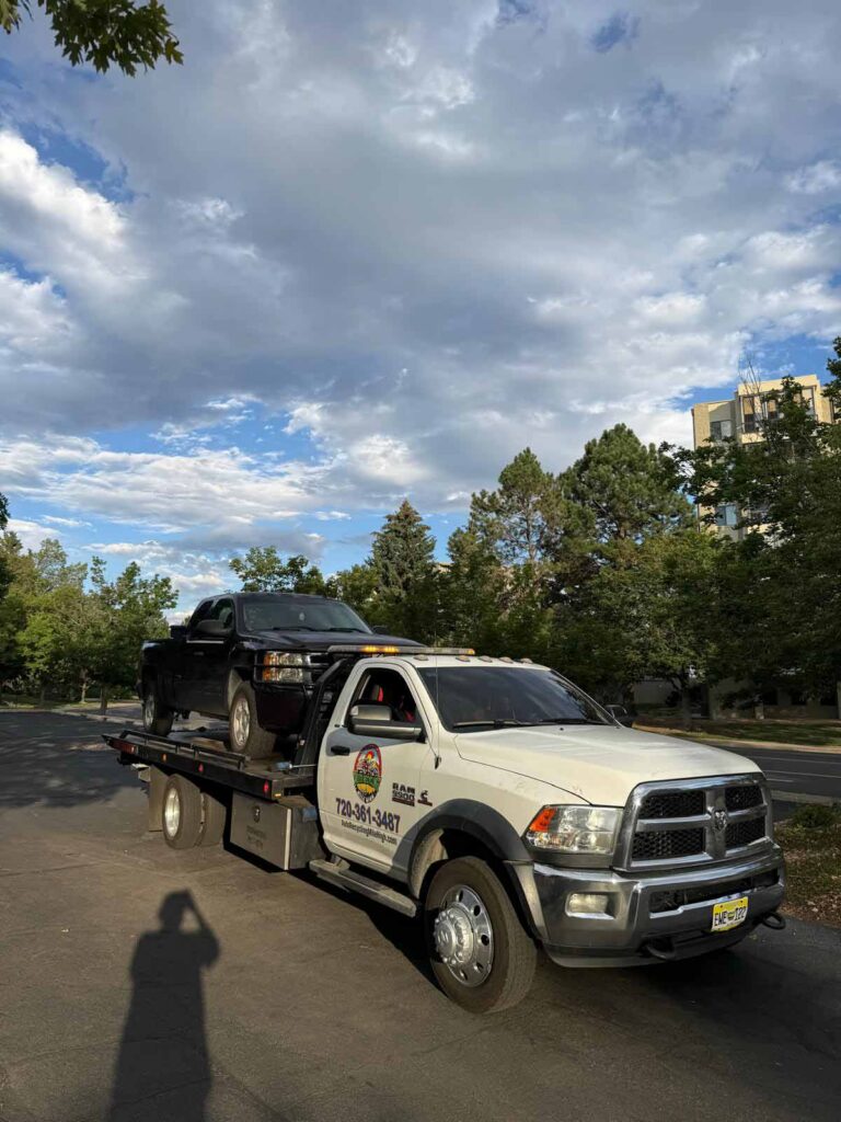 White Auto Recycling Mile High Ram 5500 flatbed with a black Chevrolet Silverado pickup strapped on the deck, parked under a dramatic cloudy Colorado sky